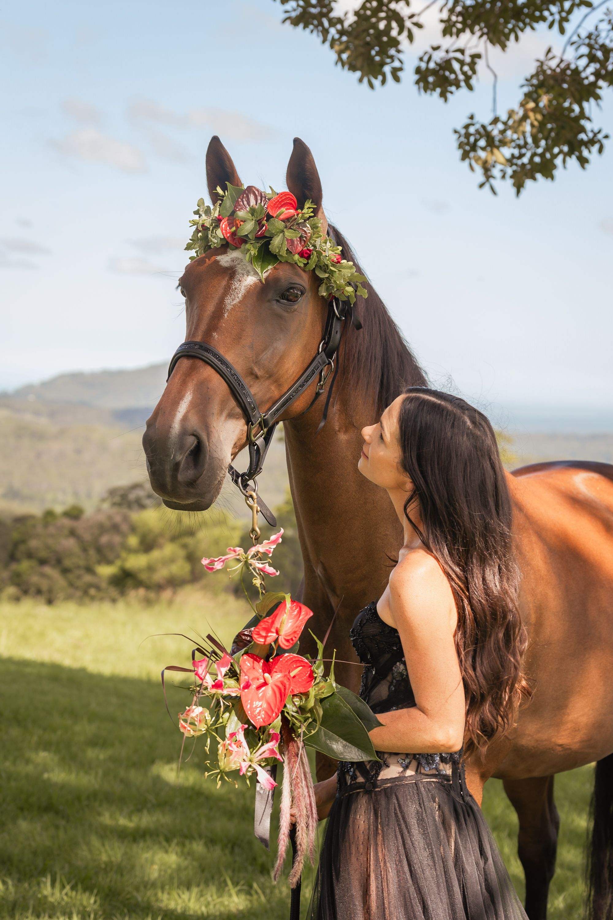 A brown horse adorned with pink floral decorations stands in a sunny meadow against a mountain backdrop.