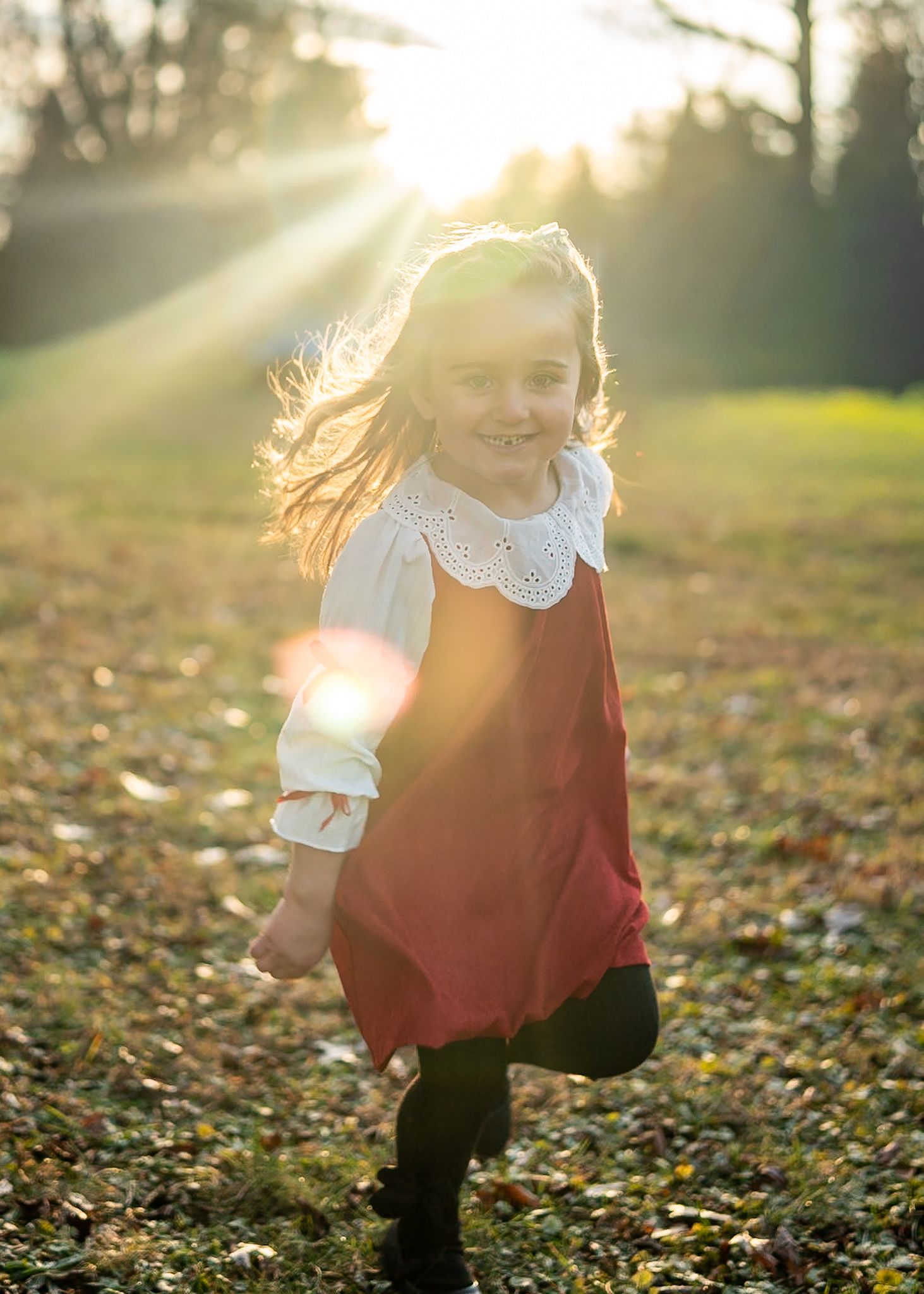 Child in a red dress playing in autumn leaves at sunset with golden sunlight streaming through trees.