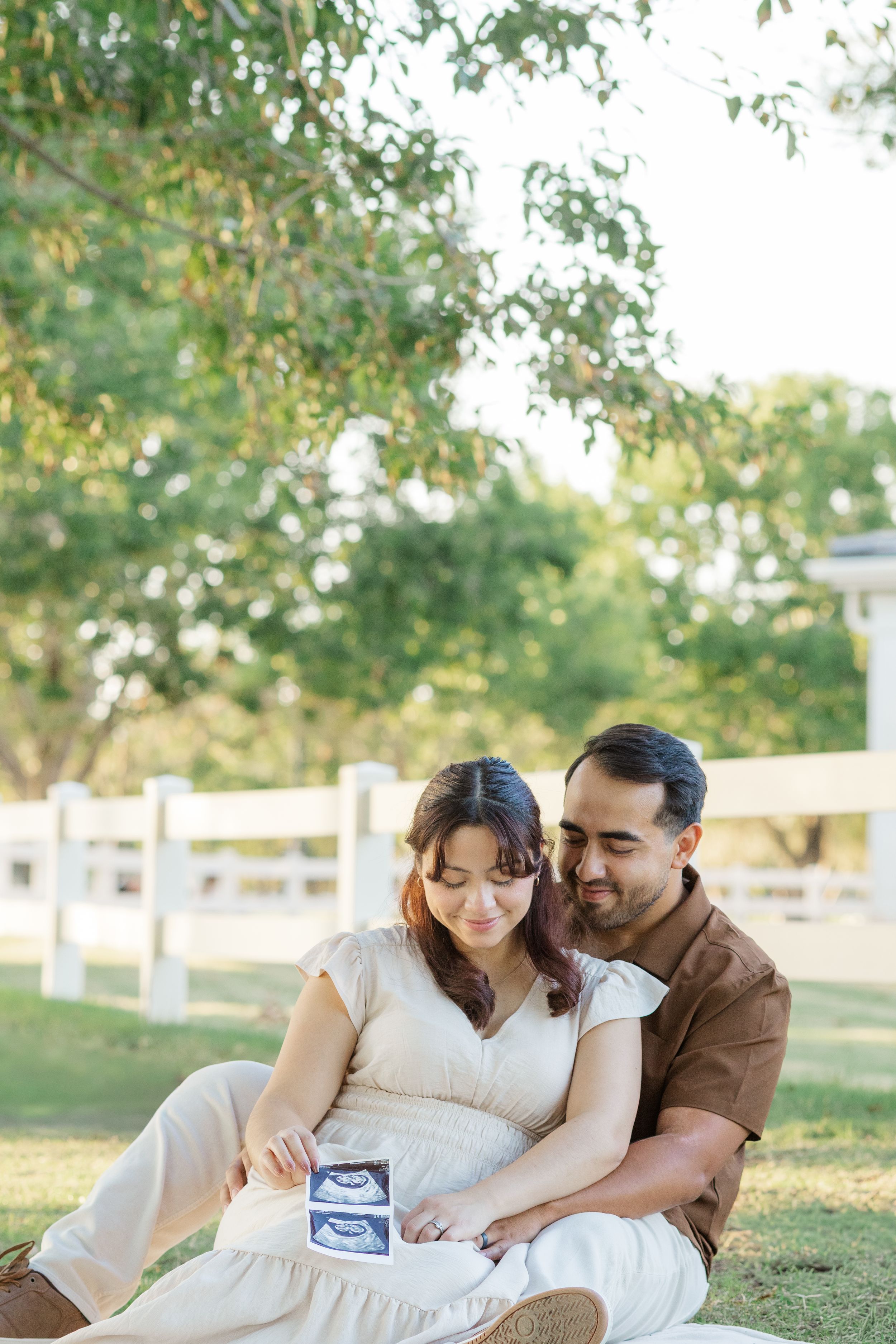Image of a young, married couple sitting on green grass in front of a white fence looking down at an ultrasound in Gilbert, Arizona.