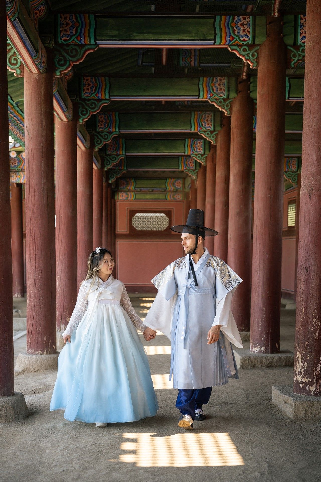 People wearing traditional white Korean hanbok clothing walk through a covered walkway with red wooden pillars at a palace.