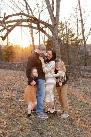 Family shares playful moments in autumn woods during golden hour sunset with fallen leaves on ground.
