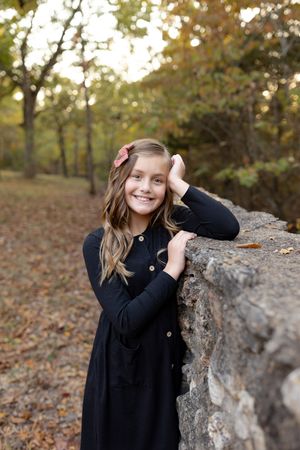 A young person in a black dress poses outdoors by a rock formation during autumn with fallen leaves in the background.