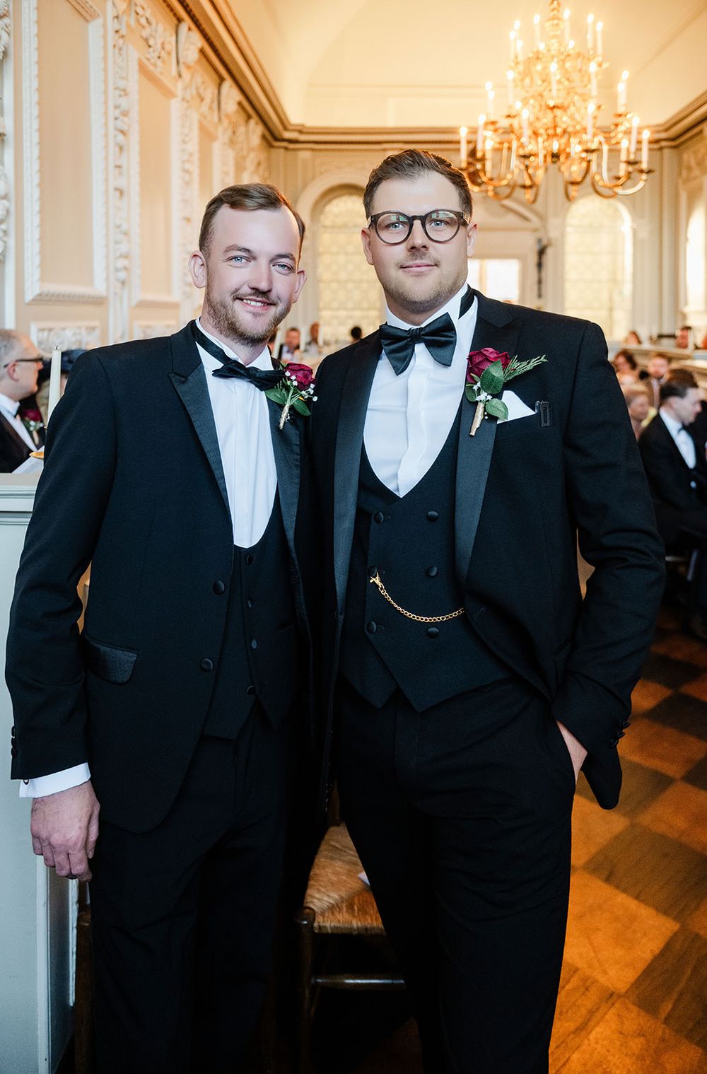 Groomsmen in black tuxedos and bowties smile together in an elegant room with chandeliers and hardwood floors.