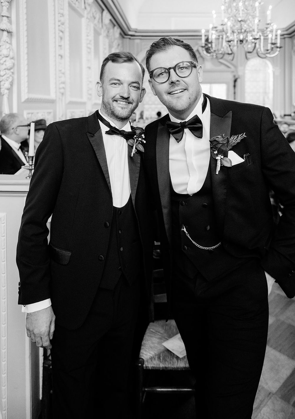 A group of groomsmen in black tuxedos and bow ties pose together at an elegant wedding reception with chandeliers.