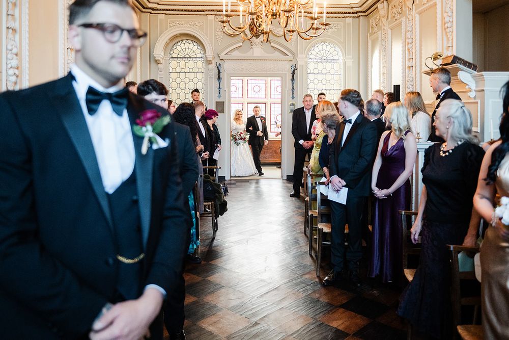 Wedding guests mingle and socialize in an elegant ballroom with crystal chandeliers and ornate white decor.