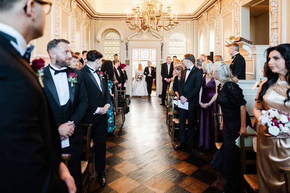 Elegant wedding ceremony in a grand ballroom with ornate chandeliers and guests watching from both sides of the aisle.