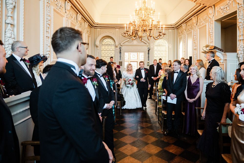 Wedding guests gather in a luxurious ballroom with black and white checkered floors and crystal chandeliers.