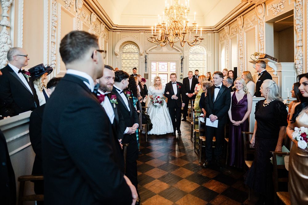 Wedding guests gather in an opulent ballroom with crystal chandeliers and herringbone wood floors during a ceremony.