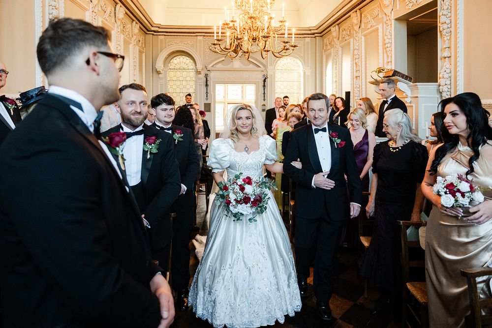 Elegant wedding celebration in an ornate ballroom with chandelier, formal attire, and floral bouquets.