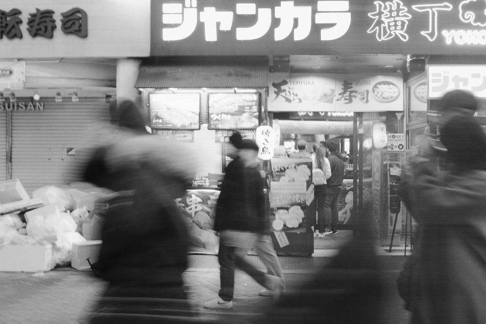 Black and white urban street scene showing blurred motion of people walking past storefronts with Japanese signage.