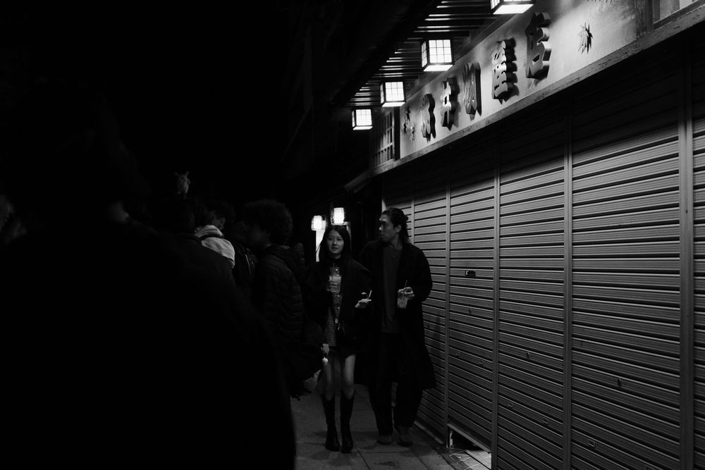 People walking through a dimly lit urban corridor with illuminated signs and metal shutters at night.