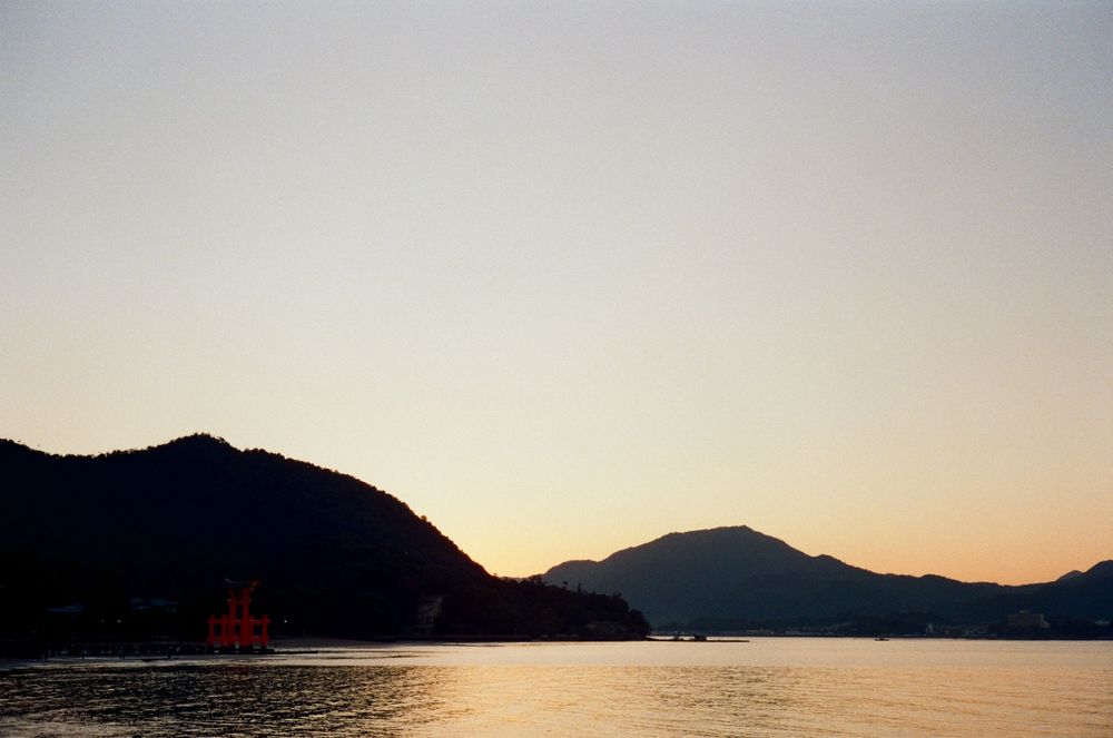 A scenic mountain landscape at sunset with silhouetted peaks against an orange and purple sky reflected in calm waters.
