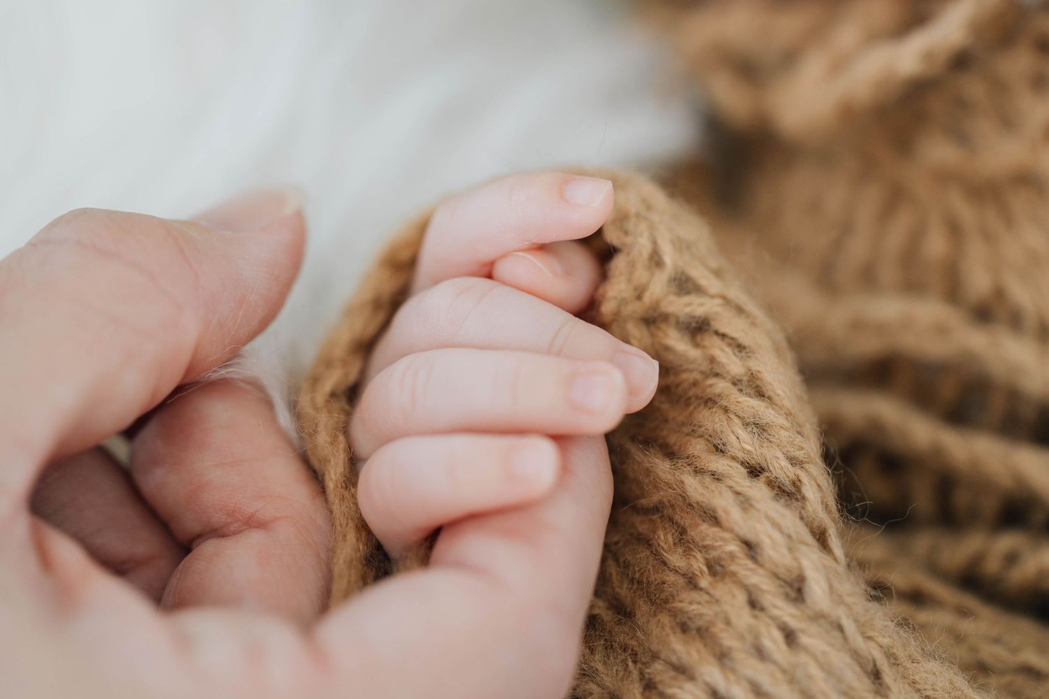 Close-up of a newborn baby hand gently grasping an adult finger on a soft brown knitted blanket.