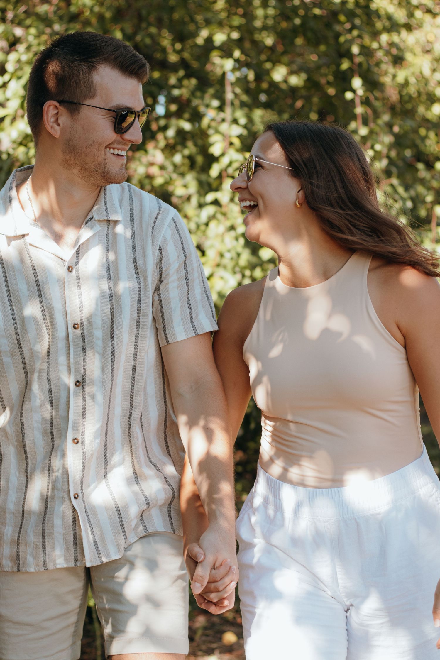 A happy couple in summer attire holding hands and laughing together in dappled sunlight.