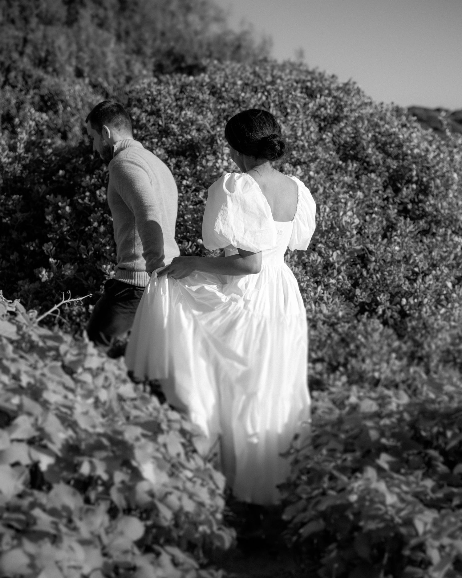 A couple walking through lush greenery wearing casual and formal attire in black and white.