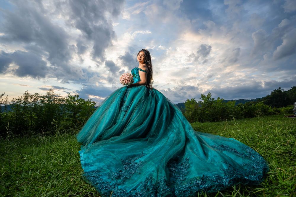 Dramatic teal ball gown photographed at sunset with cloudy sky backdrop in open field.