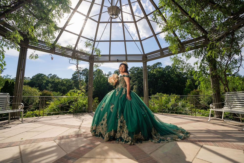 Dramatic emerald green quinceañera dress captured under an ornate glass gazebo surrounded by greenery.