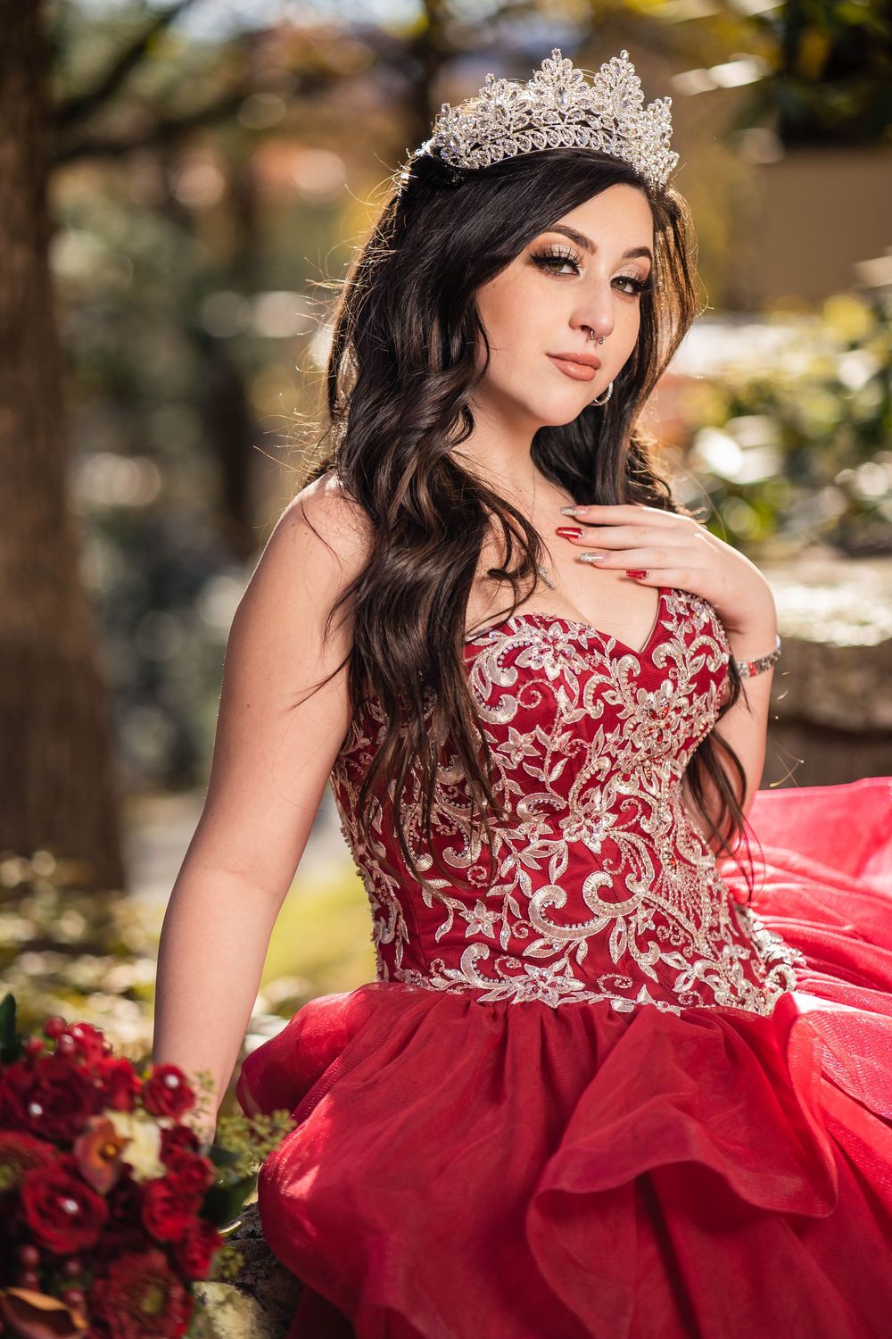 A person in a stunning red ball gown with ornate beaded bodice poses elegantly in an outdoor garden setting.