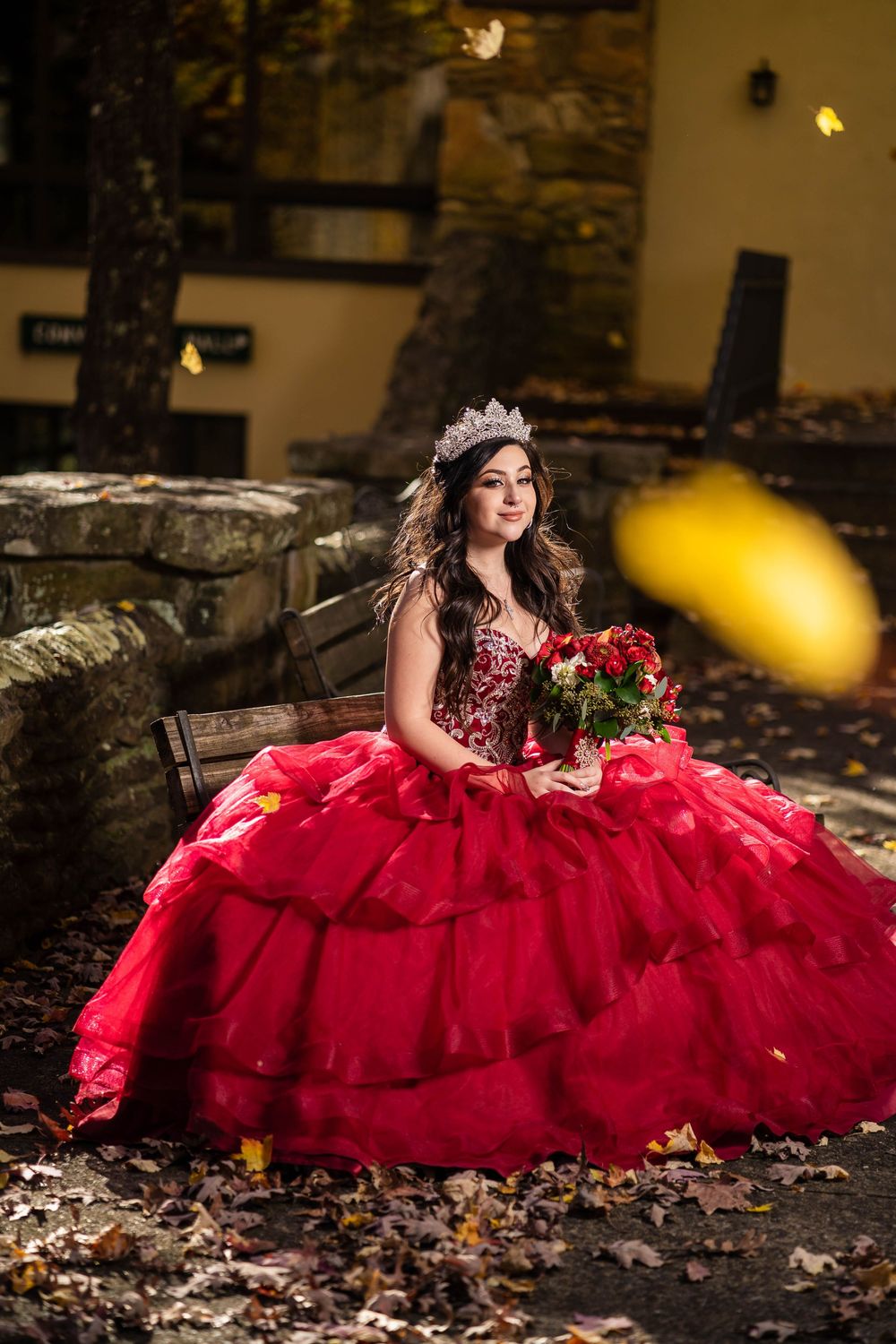 A person in a flowing red quinceañera dress poses gracefully among autumn leaves against a stone wall background.