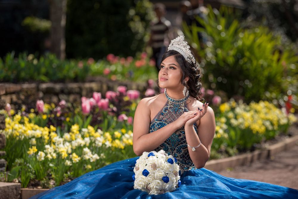 Elegant ball gown portrait in a garden with blue dress and white bouquet against colorful floral background.