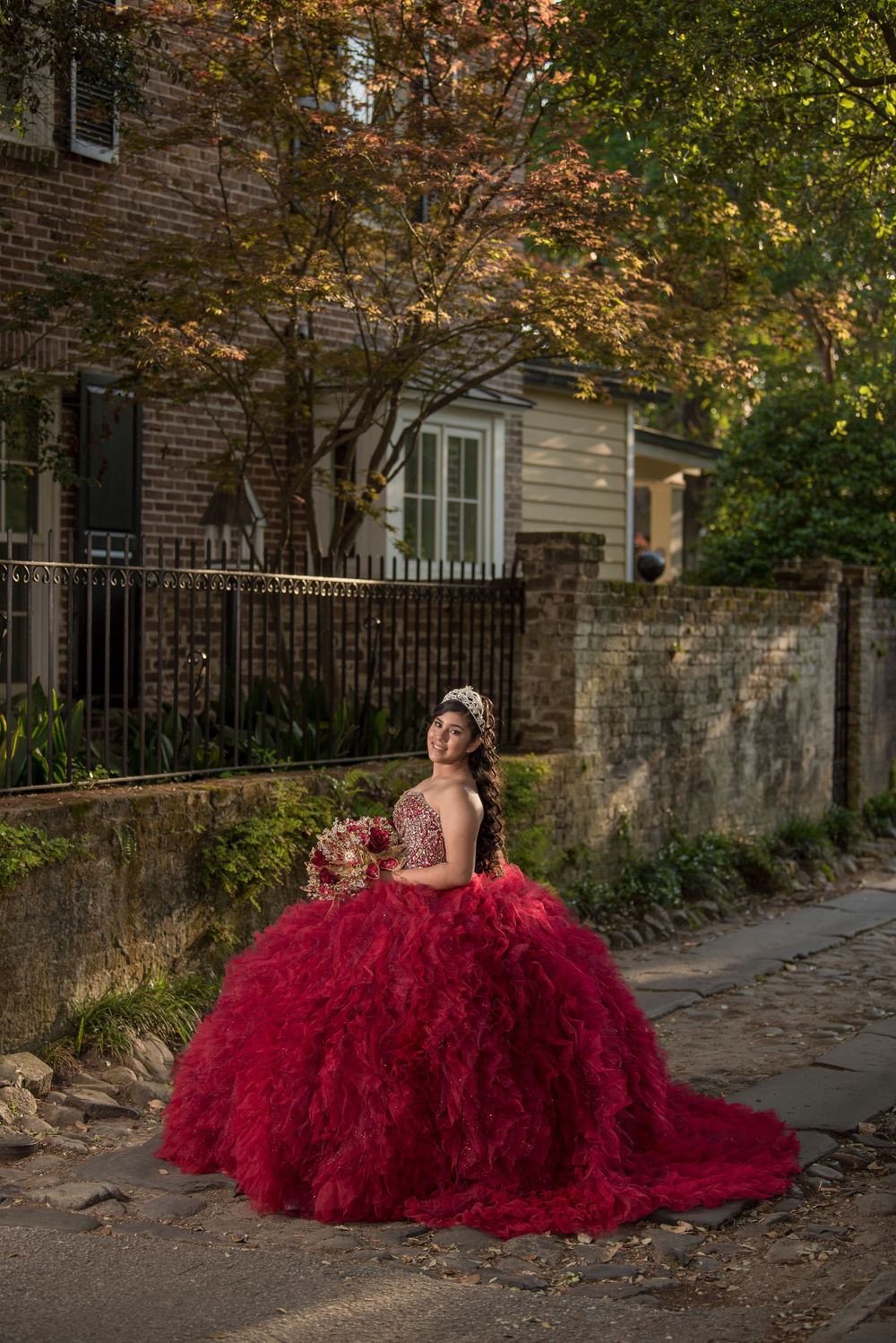 Stunning red quinceañera dress photographed on autumn street with brick wall and fallen leaves.