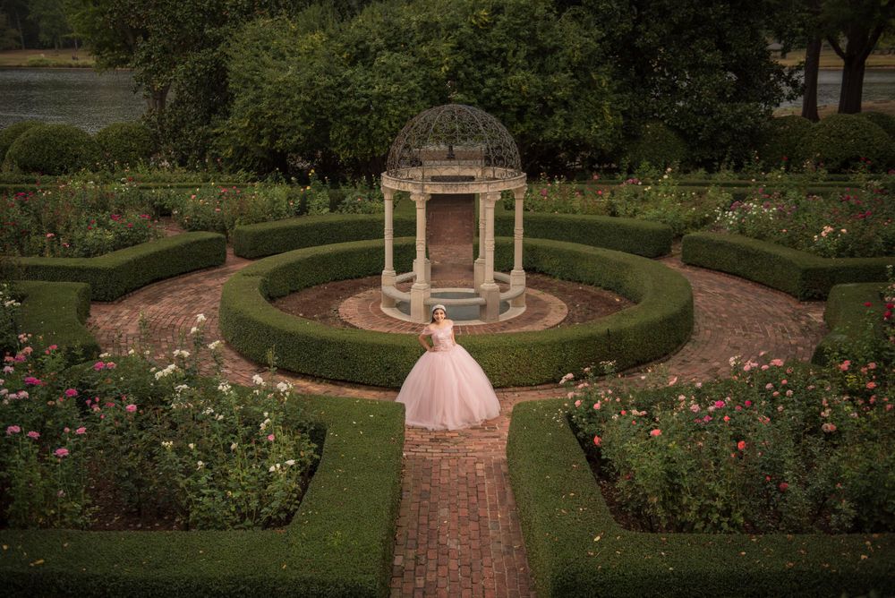Elegant pink ballgown on brick pathway leading to classical domed gazebo surrounded by manicured gardens and roses.