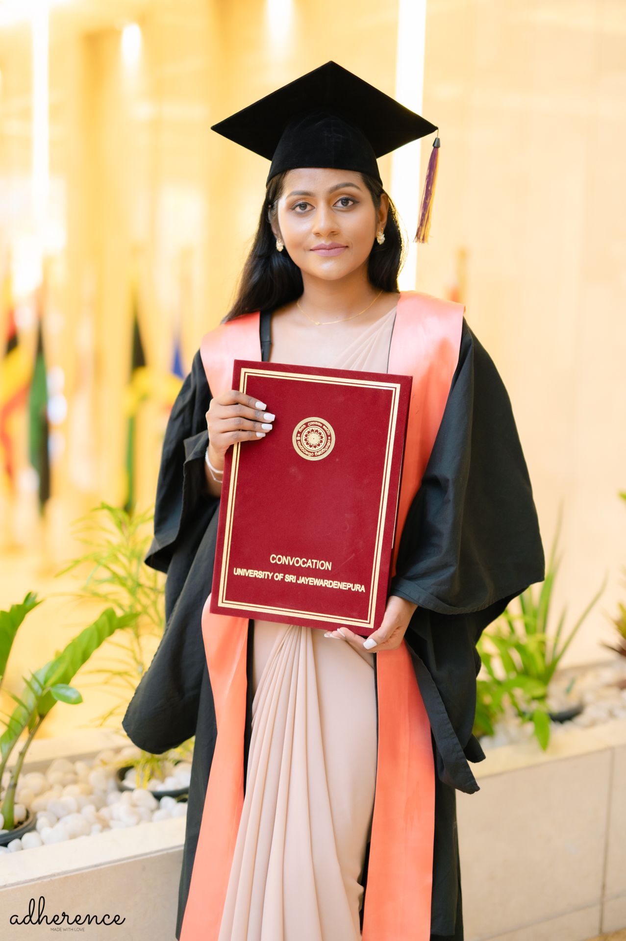 A graduate in cap and gown holds red diploma while standing against warm-lit background.
