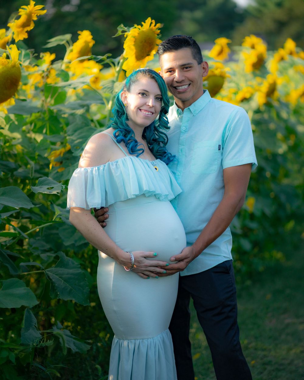 Couple posing together in dreamy maternity photoshoot among vibrant sunflower field at sunset.