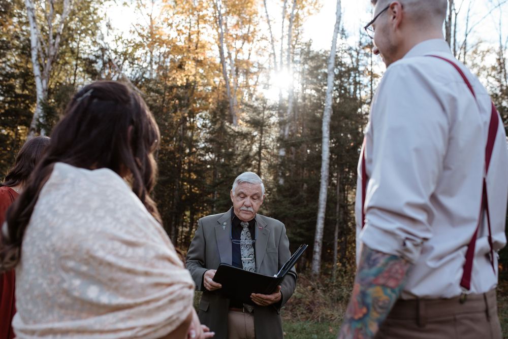 An outdoor wedding ceremony takes place in a wooded setting with attendees gathered around an officiant.