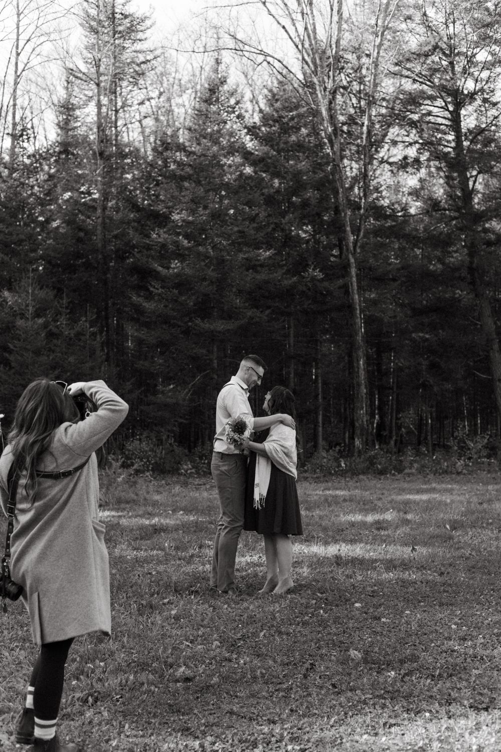 A black and white photo shows a photographer documenting an intimate moment in a forest setting.