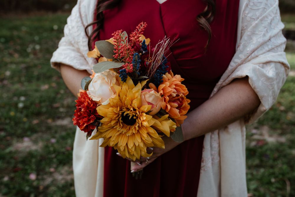 A vibrant fall bouquet with orange sunflowers and burgundy flowers held against a maroon dress.