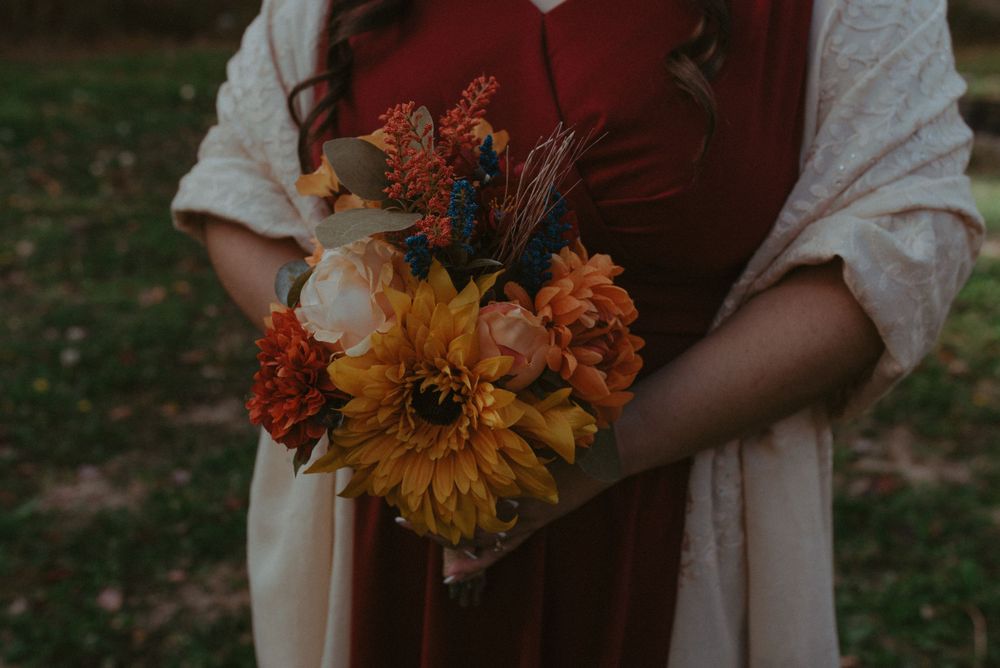 A close-up of autumn-colored bouquet featuring sunflowers and warm-toned blooms against a burgundy dress.