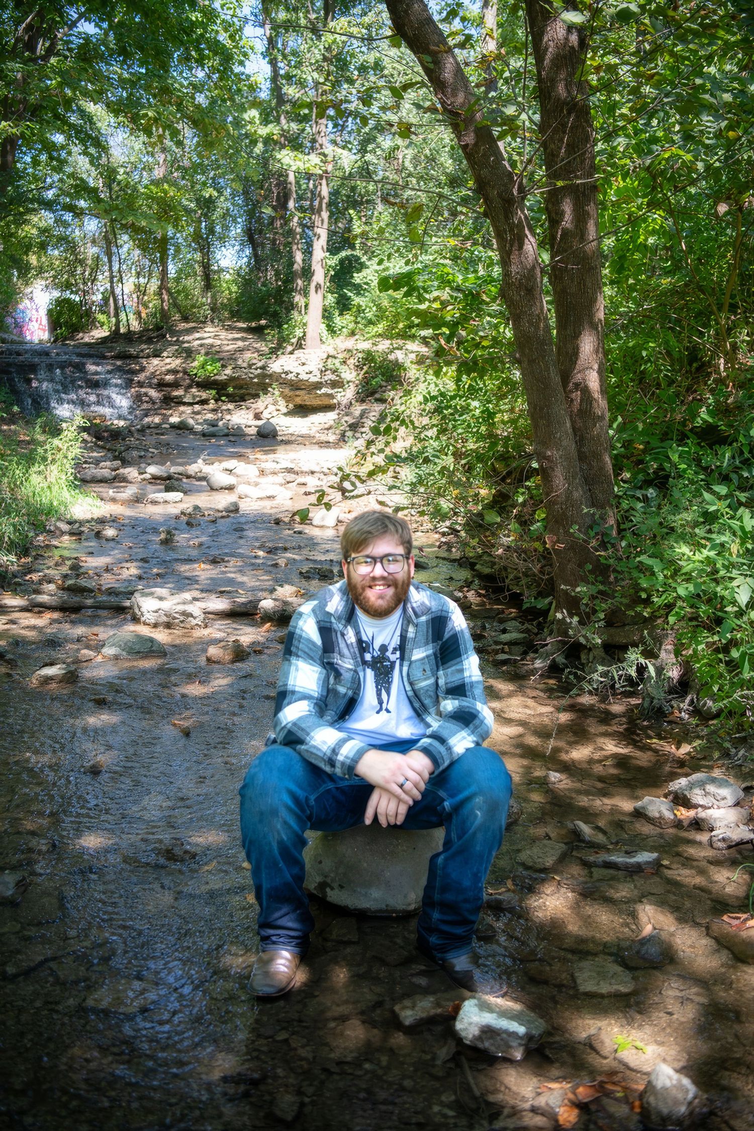 A person in plaid and denim sits on a rock along a wooded hiking trail.