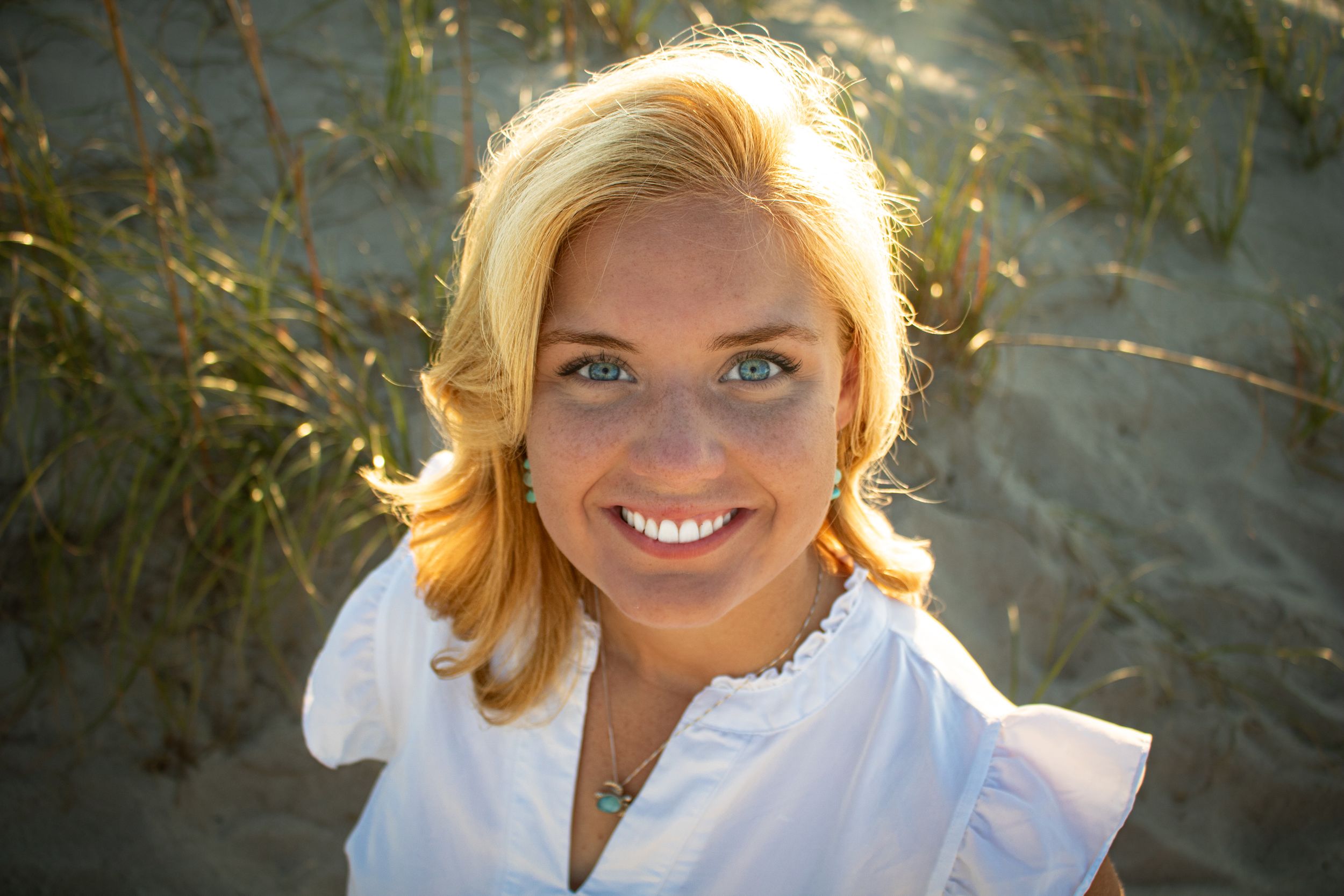 A bright smile stands out against a beach backdrop as blonde hair blows in the breeze, wearing a vibrant pink ruffled dress.