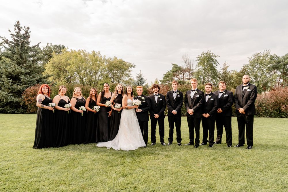 Wedding party in black dresses and suits poses together on a green lawn.