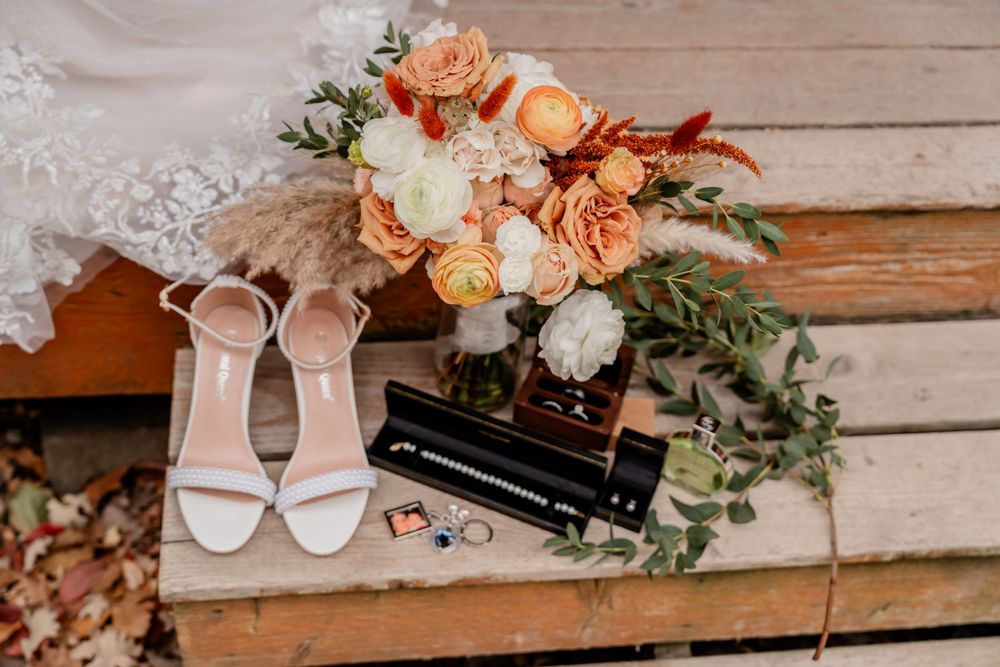Wedding details flatlay with peach flowers, white heels and rings on wooden surface.