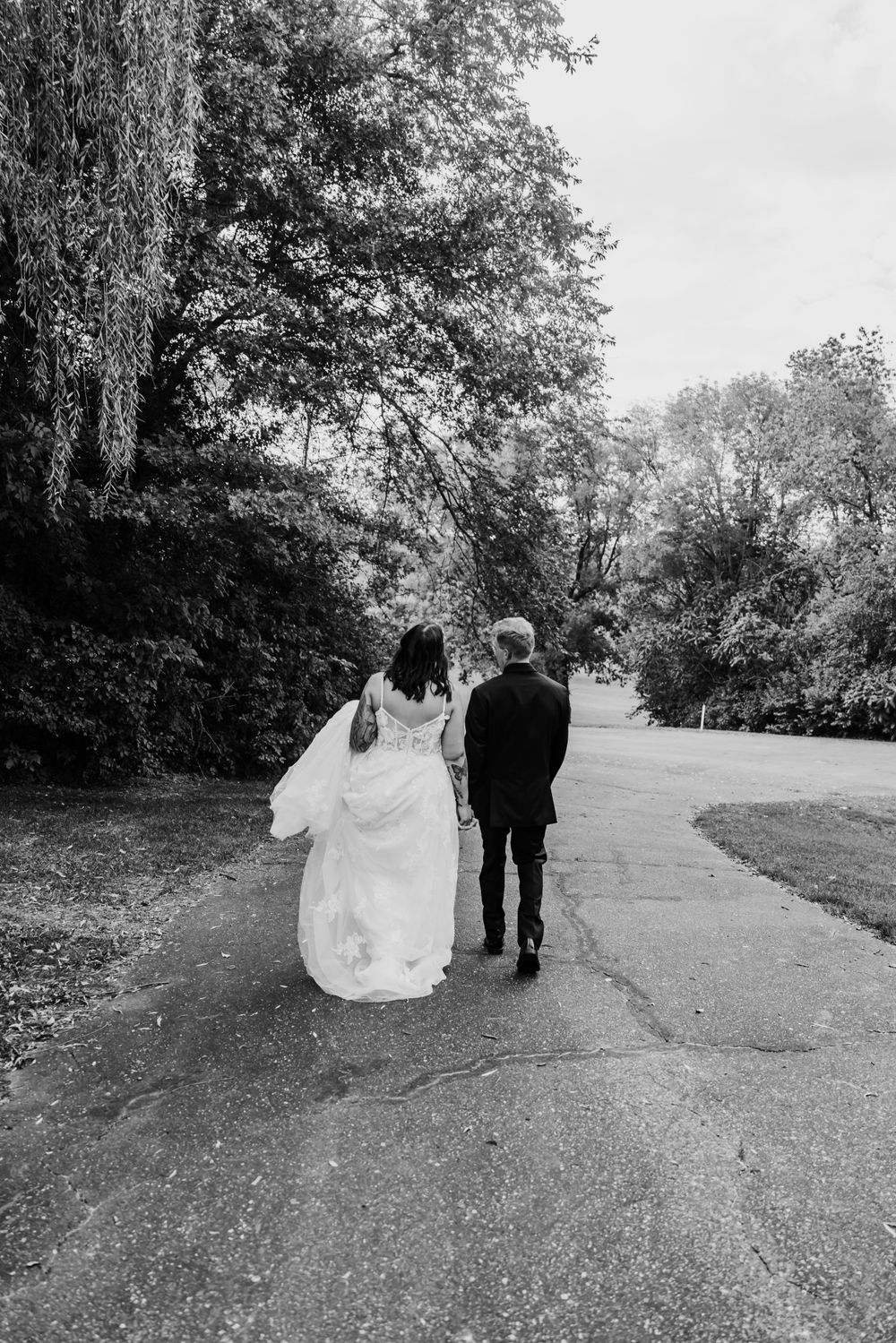 Black and white photo of newlyweds walking down a tree-lined path together.