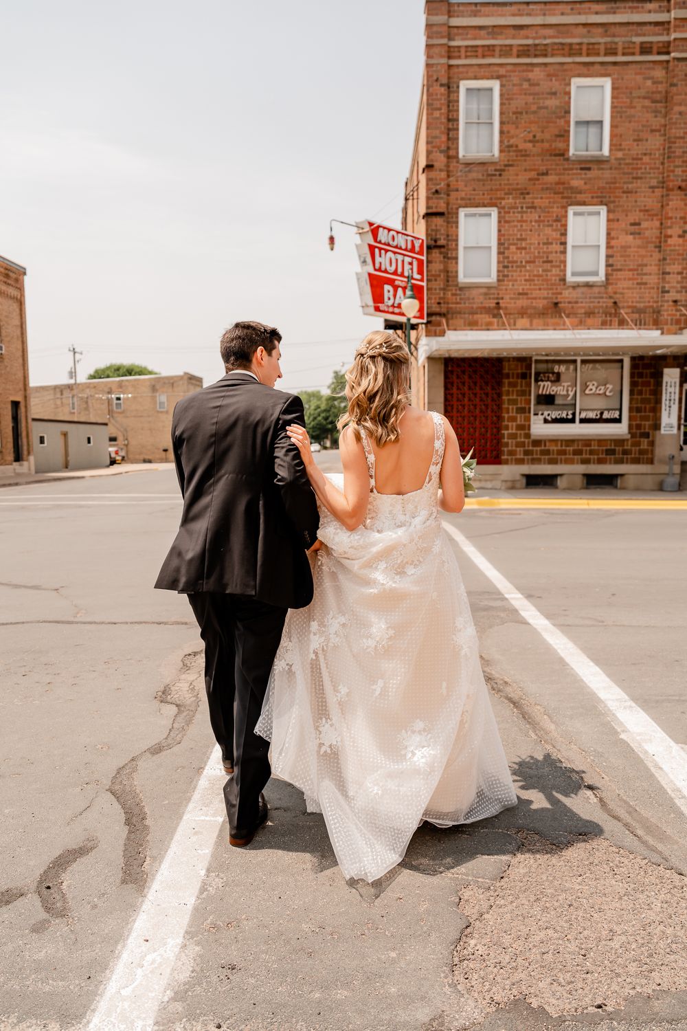 A couple in wedding attire walk down a small town street with brick buildings and vintage signs.