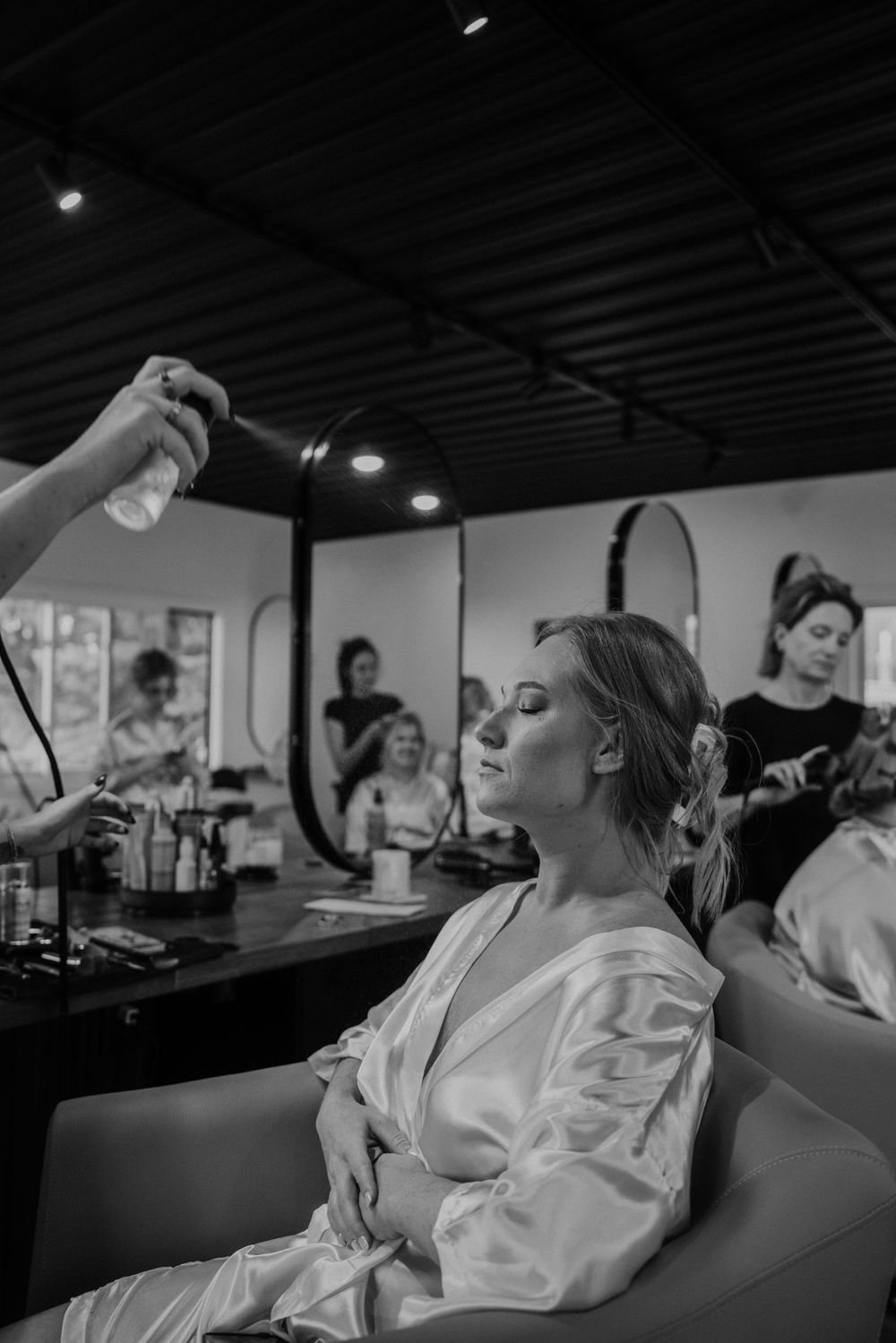 Bride getting hair styled in salon chair wearing white robe.
