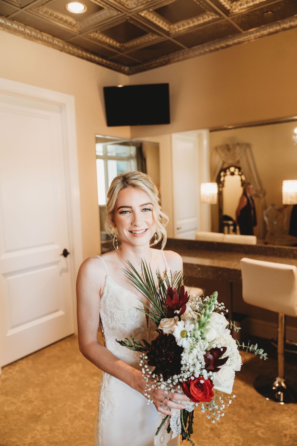 Bride smiling while holding elegant bouquet with tropical leaves and mixed flowers in getting ready room.