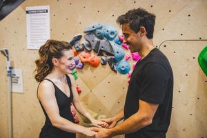 A happy couple holds hands while rock climbing at an indoor gym with colorful climbing holds on the wall at the Bouldering Project in DC