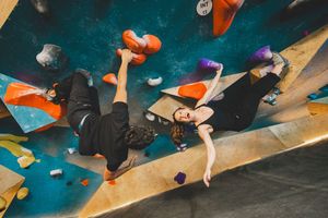 Person in black clothing climbing on an angled indoor climbing wall while holding orange dumbbells the Bouldering Project in DC