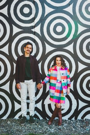 A couple poses against a black and white circular patterned wall, with one wearing a vibrant multicolored coat.