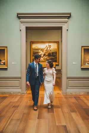 A romantic wedding couple walks through an elegant art museum gallery with hardwood floors and classical paintings on display.