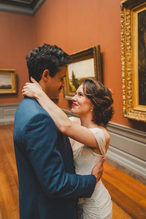 Elegant couple in wedding attire share an intimate moment while dancing in an art gallery with ornate gold frames.
