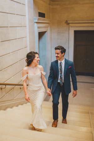 A bride in a white off-shoulder gown and groom in navy suit walk hand-in-hand up marble stairs in a grand building.