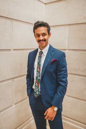 Professional wearing a navy suit with floral tie and red pocket square stands confidently against a textured wall.