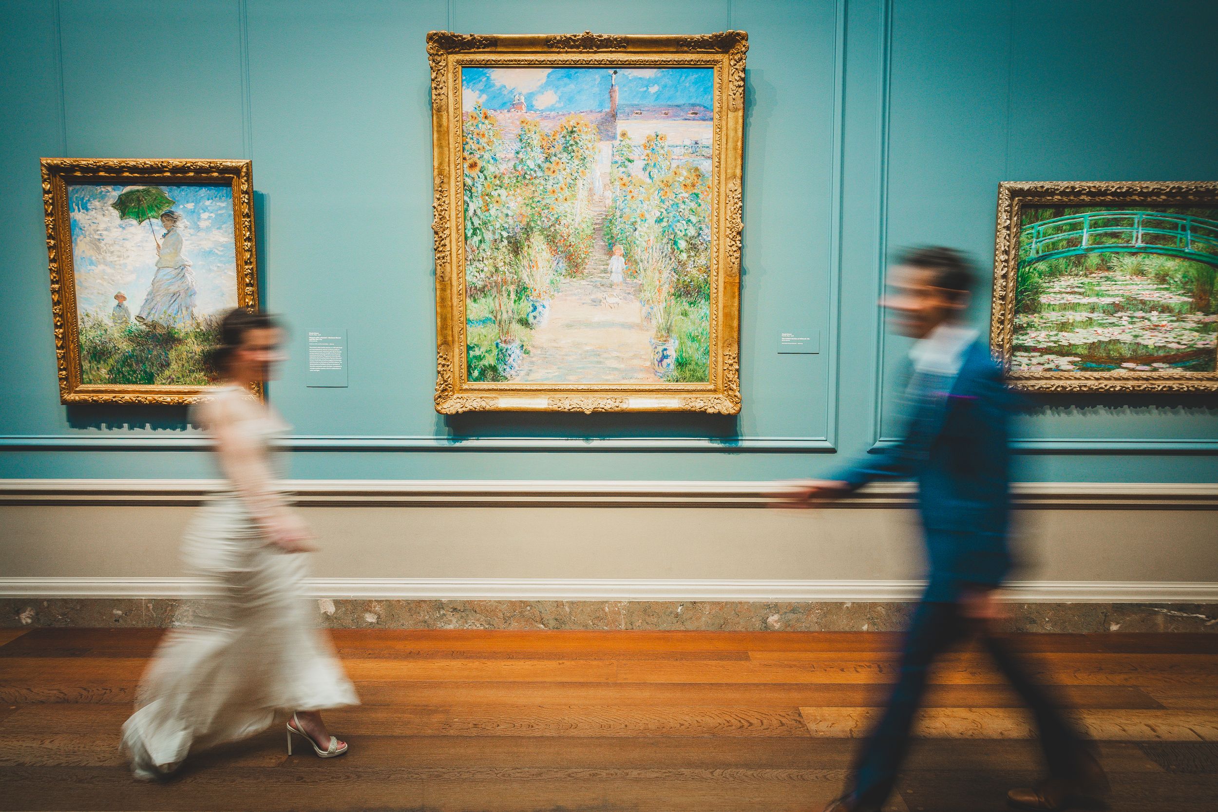 Motion-blurred visitors walk past impressionist paintings displayed on turquoise walls in an art museum gallery.