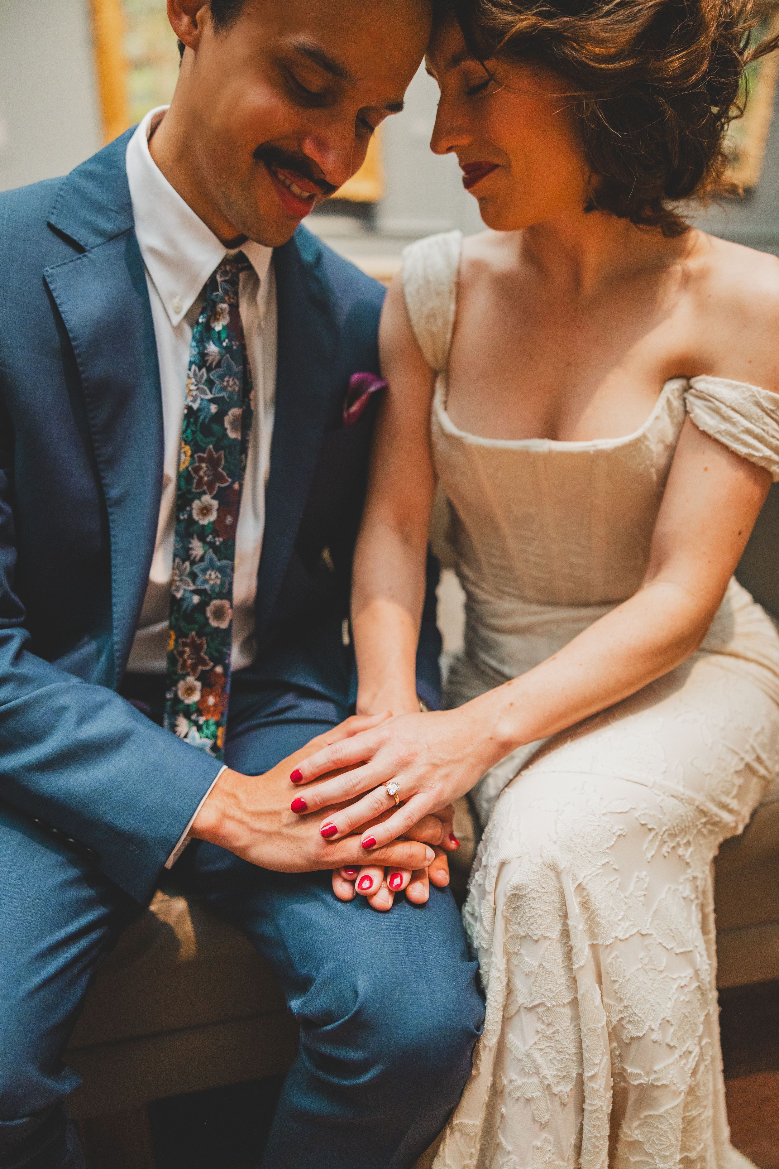 A close-up of a couple holding hands at their wedding, with him in a blue suit and her in an elegant cream dress.