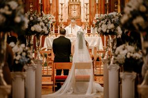 Traditional church wedding ceremony with white floral arrangements on pillars and long flowing veil down the aisle.
